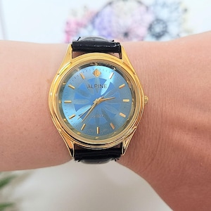 May include: A gold-toned Alpine Quartz watch with a blue face and black leather band. The watch face has a textured design and gold hands. The watch is on a wrist.