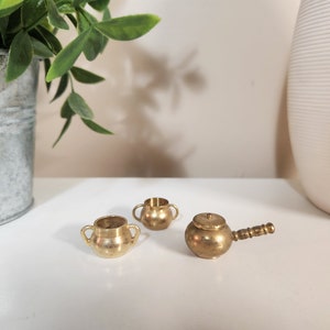 Peut inclure: Trois petits récipients en métal de couleur dorée avec des poignées. Deux sont ronds et un a un couvercle et une poignée.