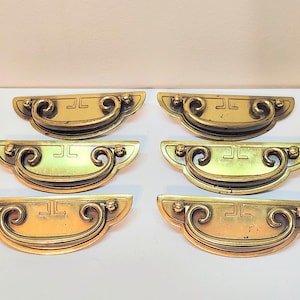 Può includere: Sei maniglie per cassetti vintage in ottone con manico curvo e dettagli decorativi. Ogni maniglia presenta un design rettangolare con un simbolo stilizzato. Le maniglie sono disposte in due file da tre, mostrando la loro finitura dorata invecchiata.