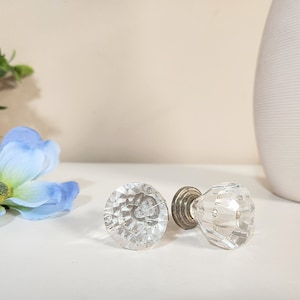 Könnte beinhalten: Zwei klare, facettierte Glasschrankknöpfe mit silberfarbenen Metallsockeln. Die Knöpfe sind rund und haben ein rautenförmiges Muster. Der Hintergrund umfasst eine blaue Blume, eine kleine grüne Pflanze und eine weiße Vase.