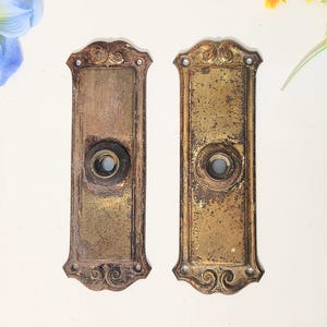 May include: Two antique brass door plates with ornate detailing. Each plate has a central hole for a doorknob and decorative scrollwork at the top and bottom. The plates have a weathered, aged appearance, with visible wear and discoloration.