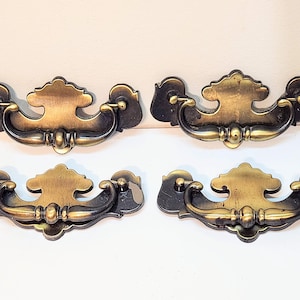以下が含まれることがあります： 装飾的なデザインの、真鍮製のアンティーク引き出し取っ手4つ。各取っ手は、湾曲したハンドルと装飾的なバックプレートが特徴です。真鍮は暖かく、古びた風合いで、ヴィンテージ感を加えています。家具の修復プロジェクトに最適です。