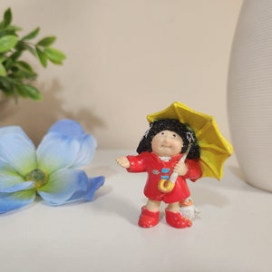 May include: A small plastic figurine of a girl with black hair wearing a red coat and holding a yellow umbrella.