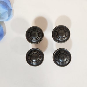 May include: Four black metal cabinet knobs with a circular design and a raised centre. The knobs have a distressed finish.