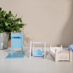 May include: A set of three miniature furniture pieces for a dollhouse. The set includes a blue swing set, a blue shelf unit, and a white bed with pink floral details.