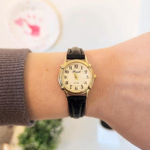 May include: A gold-tone wristwatch with a black leather band. The watch face is white with black Roman numeral hour markers and black hands. The watch face also has the word "Royale" and "Quartz" printed on it.