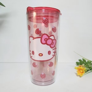 Op de afbeelding: Een doorzichtige, dubbelwandige beker met een rode deksel. De beker heeft een roze Hello Kitty-afbeelding en verspreide aardbeienontwerpen. Ontworpen voor koude dranken.