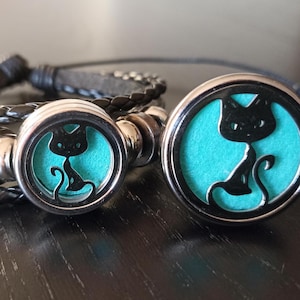 May include: A black leather bracelet with a silver charm featuring a black cat silhouette on a turquoise background. The bracelet has a braided design and is displayed on a dark surface. The charm is round and has a metallic border.