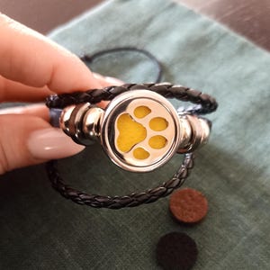 May include: A black leather bracelet with a silver charm featuring a yellow paw print. The charm has a small opening for adding essential oils.