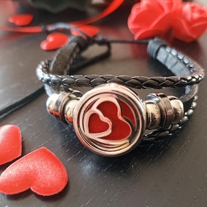May include: A black leather bracelet with a silver charm featuring two red hearts. The bracelet is layered with braided leather and has silver beads.