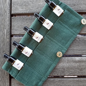 Puede incluir: Un soporte de lino verde para aceites esenciales con cinco botellas de vidrio pequeñas. Cada botella tiene un cuentagotas negro y una etiqueta blanca con las palabras "Nature Arc" y el nombre del aceite esencial. El soporte se asegura con botones de madera.