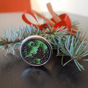 May include: A silver diffuser with a green insert featuring a stylised Christmas tree design. The diffuser is sitting on a dark wood surface with a pine branch.
