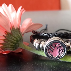 May include: A black leather bracelet with a silver charm featuring a pink angel wing design. The bracelet is layered with a braided leather strap.