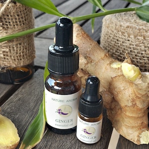 Puede incluir: Dos botellas de vidrio ámbar con goteros llenas de aceite esencial de jengibre. Las botellas tienen etiquetas blancas con el texto "Nature Aroma" y "Aceite esencial de jengibre". Se puede ver un trozo de raíz de jengibre en el fondo.