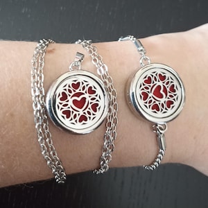 Op de afbeelding: Een zilverkleurige kettinghanger en een bijpassende armband, beide met een rond medaillonontwerp met een ingewikkeld filigraanpatroon van hartjes op een effen rode achtergrond. De sieraden zijn verbonden aan zilverkleurige schakelkettingen.
