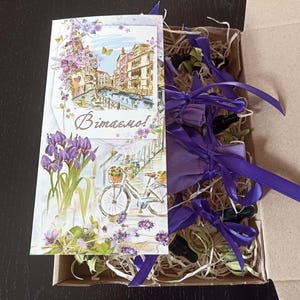 May include: A gift box filled with lavender sachets tied with purple ribbon. The box is decorated with a card featuring a watercolour illustration of a cityscape with a bicycle and the word "Vitamo!" in a cursive font.
