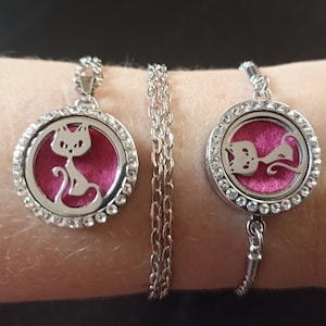 May include: A silver-toned necklace and bracelet set. Each piece features a round pendant with a cat silhouette on a pink background, surrounded by clear crystals. The necklace has a chain, and the bracelet has a chain and a charm.
