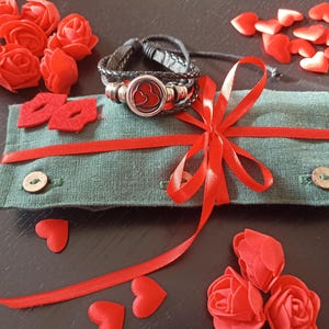 May include: A green fabric gift bag with a red ribbon and bow. The bag contains a black leather bracelet with a silver heart charm. The bag is surrounded by red felt hearts.