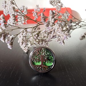 May include: A silver aromatherapy diffuser featuring a tree of life design and a green interior. The diffuser is placed on a dark surface, with dried lavender-coloured flowers in the background.
