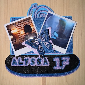 May include: A blue and purple cake topper featuring a butterfly design. The topper includes two photographs, one of a person and one of a landscape. The topper also displays the name "Alyssa" and the number "13".