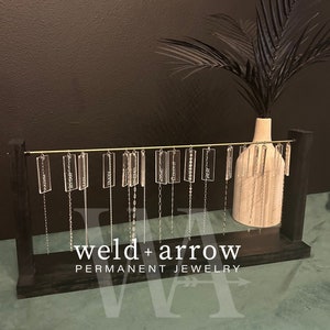 May include: A black wooden display stand with a gold rod holding multiple silver chains with clear acrylic tags. Each tag has a different word engraved on it. The stand has the text "weld + arrow PERMANENT JEWELRY" on the front.