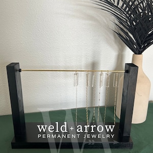 May include: A black wooden jewelry display stand with a gold metal bar and six clear glass pendants hanging from it. The stand has the text "weld+arrow PERMANENT JEWELRY" on the front.