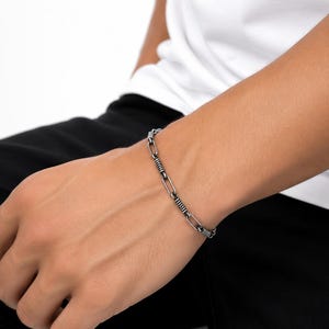 May include: A silver-toned metal chain bracelet featuring alternating elongated oval links and smaller, textured cylindrical segments with a dark, oxidized finish, creating a distinct industrial appearance on the wrist.