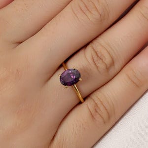 May include: A gold ring featuring an oval, deep purple gemstone. The ring's band is thin and simple, with the gemstone held in place by four prongs. The ring is shown on a hand with red nail polish.