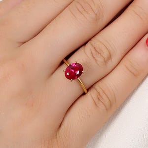 May include: A gold ring featuring a large, oval-cut, deep red gemstone. The ring's band is a thin, polished gold, and the gemstone is set with small prongs. The ring is displayed on a hand with red nail polish.
