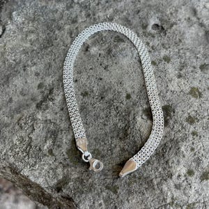 May include: A silver bracelet with a mesh design, resting on a gray stone surface. The bracelet is made of small, interconnected links, creating a flexible, textured appearance. The clasp is visible, and the bracelet is unclasped.