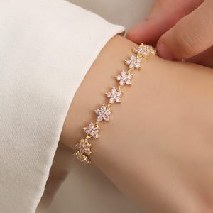 May include: A gold-colored bracelet featuring a series of flower-shaped links. Each flower has small, pink-colored accents. The bracelet is worn on a wrist, and the clasp is not visible. The background is a white shirt.