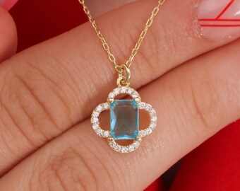 May include: A gold-colored necklace with a pendant featuring a rectangular, light blue gemstone surrounded by small, clear stones in a four-leaf clover design. The necklace is displayed against a red background.