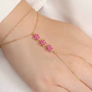 Könnte beinhalten: Eine goldfarbene Handkette mit drei rosa blütenförmigen Akzenten. Die filigrane Kette verbindet sich mit einem Ring und umschließt das Handgelenk. Der Schmuck wird an einer Hand mit weißem Hintergrund getragen.