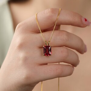 Può includere: Una collana a catena dorata con un ciondolo in pietra preziosa rosso scuro, rettangolare e sfaccettato, fissato da una montatura a quattro griffe. Il ciondolo pende da una delicata catena a maglie dorate, mostrando una pietra rossa vivace.