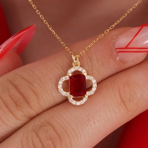 Może przedstawiać: Złoty naszyjnik z zawieszką w kształcie czterolistnej koniczyny. Zawieszka ma prostokątny, głęboko czerwony kamień szlachetny pośrodku, otoczony małymi, przezroczystymi kamieniami. Na czerwonym tle.