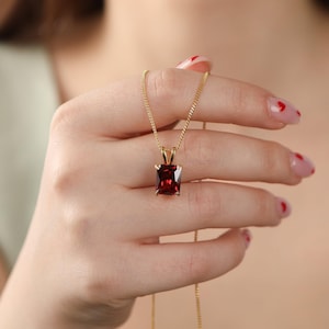 Puede incluir: Un collar de cadena de oro con un colgante de forma cuadrada que presenta una gran piedra roja oscura.