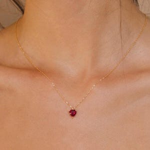 Op de afbeelding: Een delicate gouden ketting met een kleine, gefacetteerde hartvormige hanger van rode edelsteen. De ketting rust op het sleutelbeen en toont een dunne, gepolijste metalen schakelketting die de levendige karmozijnrode hartvormige bedel vasthoudt.