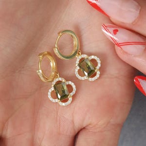 Peut inclure: Boucles d'oreilles créoles dorées, chacune ornée d'un motif de trèfle à quatre feuilles. Le trèfle est orné d'une pierre verte au centre et entouré de petits cristaux transparents. Présentées sur un fond clair.