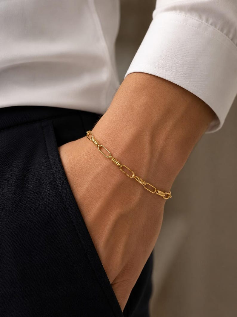 May include: A gold-toned chain bracelet featuring a repeating pattern of elongated oval links alternating with smaller, textured cylindrical segments. The bracelet has a polished finish and a consistent link design throughout its entire length.