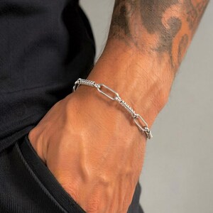 May include: A silver-toned chain bracelet featuring alternating links. The design consists of smooth, elongated oval links connected by smaller, textured cylindrical links with a ribbed surface pattern, creating a distinct metallic appearance.