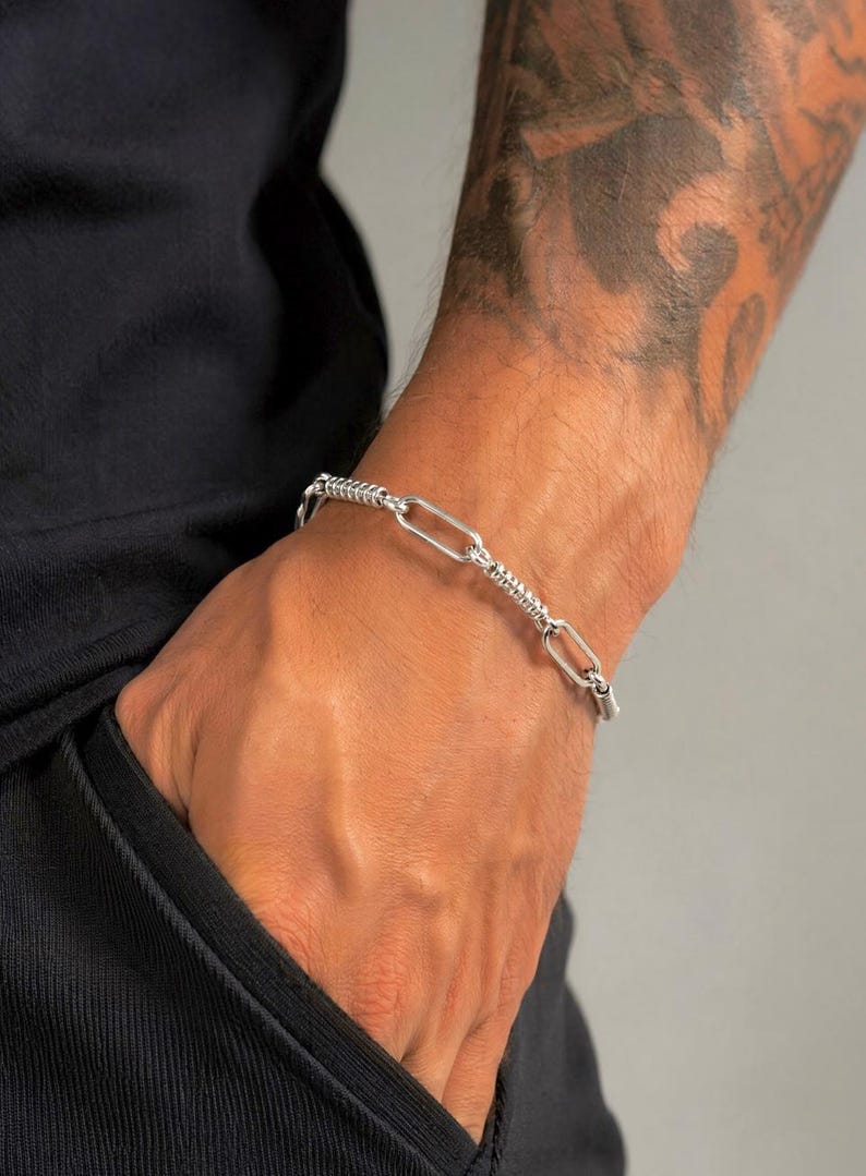 May include: A silver-toned chain bracelet featuring alternating links. The design consists of smooth, elongated oval links connected by smaller, textured cylindrical links with a ribbed surface pattern, creating a distinct metallic appearance.
