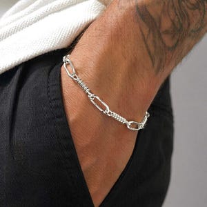 May include: A silver-toned chain bracelet featuring a repeating pattern of elongated oval links alternating with sections of tightly coiled, textured wire. The metallic links are polished and reflective, creating a continuous, flexible band around the wrist.