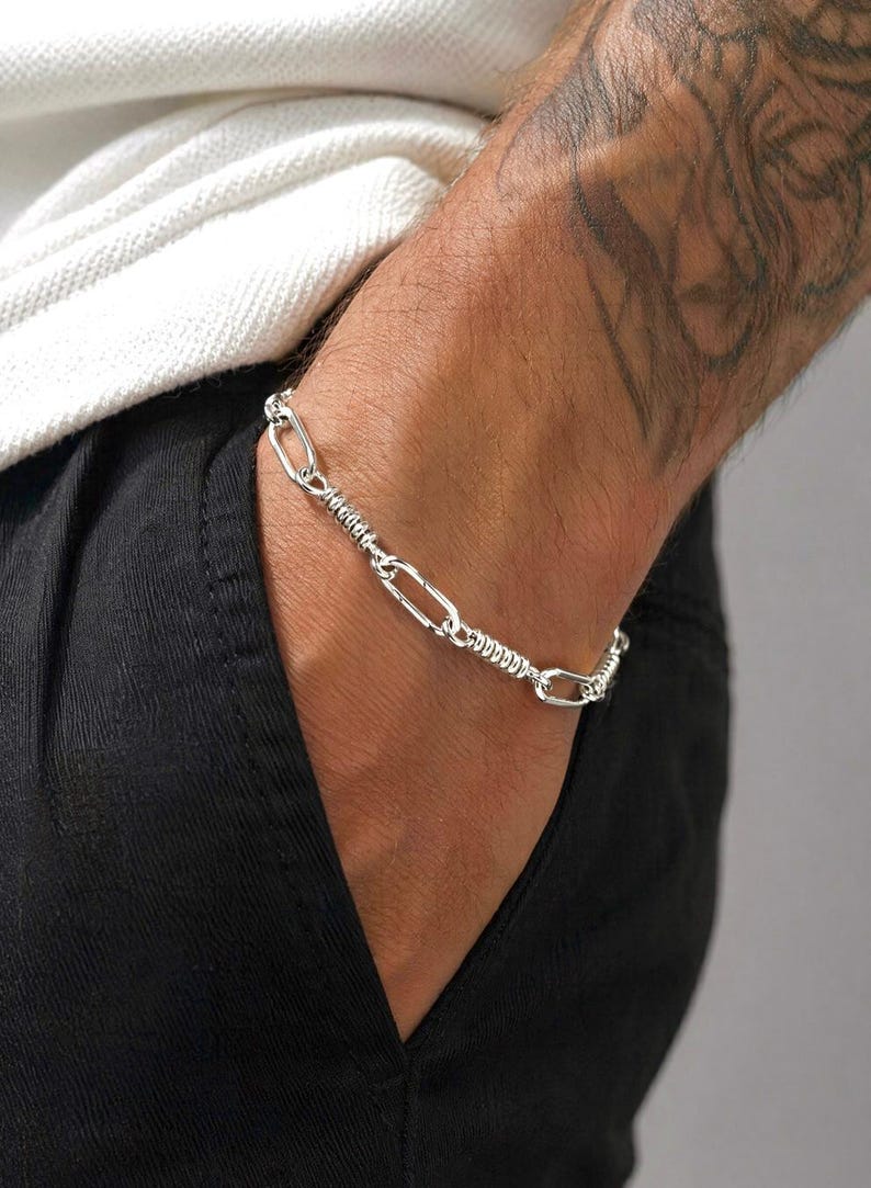 May include: A silver-toned chain bracelet featuring a repeating pattern of elongated oval links alternating with sections of tightly coiled, textured wire. The metallic links are polished and reflective, creating a continuous, flexible band around the wrist.