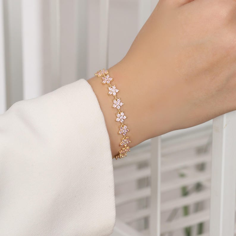 May include: A gold-colored bracelet featuring a floral design with pink and clear gemstones. The bracelet is worn on a wrist, showcasing the delicate details of the jewelry. The flowers are evenly spaced along the chain.