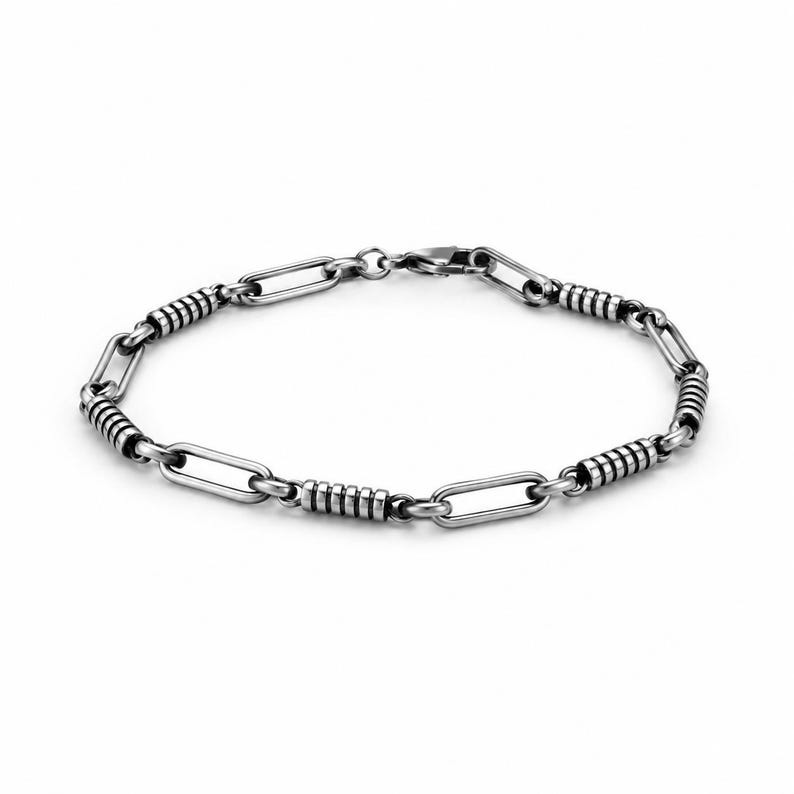 May include: A silver-toned metal chain bracelet featuring alternating elongated oval links and cylindrical segments with horizontal grooved detailing. The bracelet is secured with a small lobster claw clasp, showcasing a polished finish with darkened accents in the grooves.