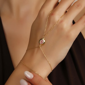 May include: A gold-tone hand chain bracelet featuring a delicate chain that connects a wrist band to a central ring component. The central piece is a gold-tone pomegranate-shaped charm inlaid with small, sparkling pink stones at its center.