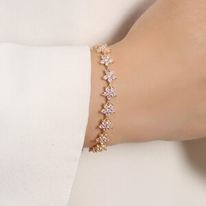 May include: A gold-colored bracelet featuring a series of delicate flower-shaped charms. Each charm has a central pink stone surrounded by smaller clear stones. The bracelet is worn on a wrist against a white background.