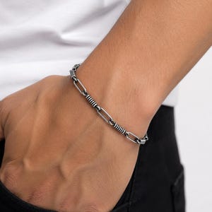 May include: A silver-toned metal chain bracelet featuring alternating elongated oval links and smaller cylindrical segments with a ribbed, textured finish. The bracelet has a polished, dark metallic appearance and a secure clasp mechanism.
