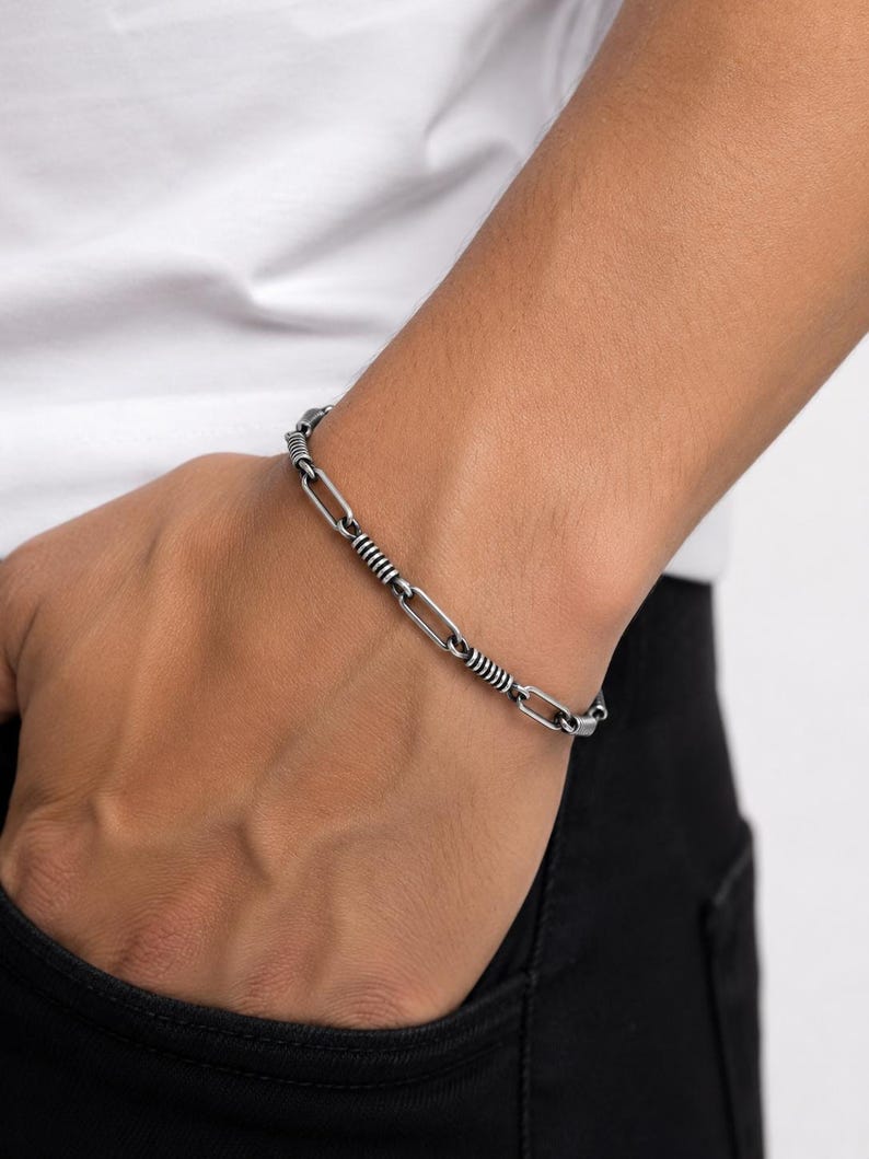 May include: A silver-toned metal chain bracelet featuring alternating elongated oval links and smaller cylindrical segments with a ribbed, textured finish. The bracelet has a polished, dark metallic appearance and a secure clasp mechanism.