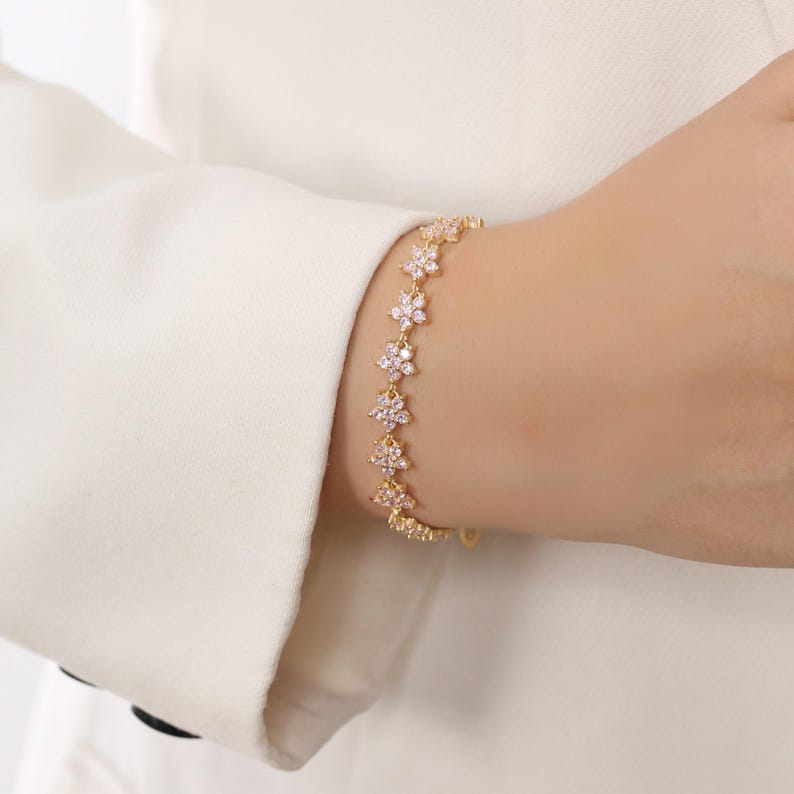 May include: A gold-toned bracelet featuring a series of delicate, flower-shaped links. Each flower is adorned with small, sparkling crystals, creating a subtle, elegant shimmer. The bracelet is worn on a wrist, showcasing its design.